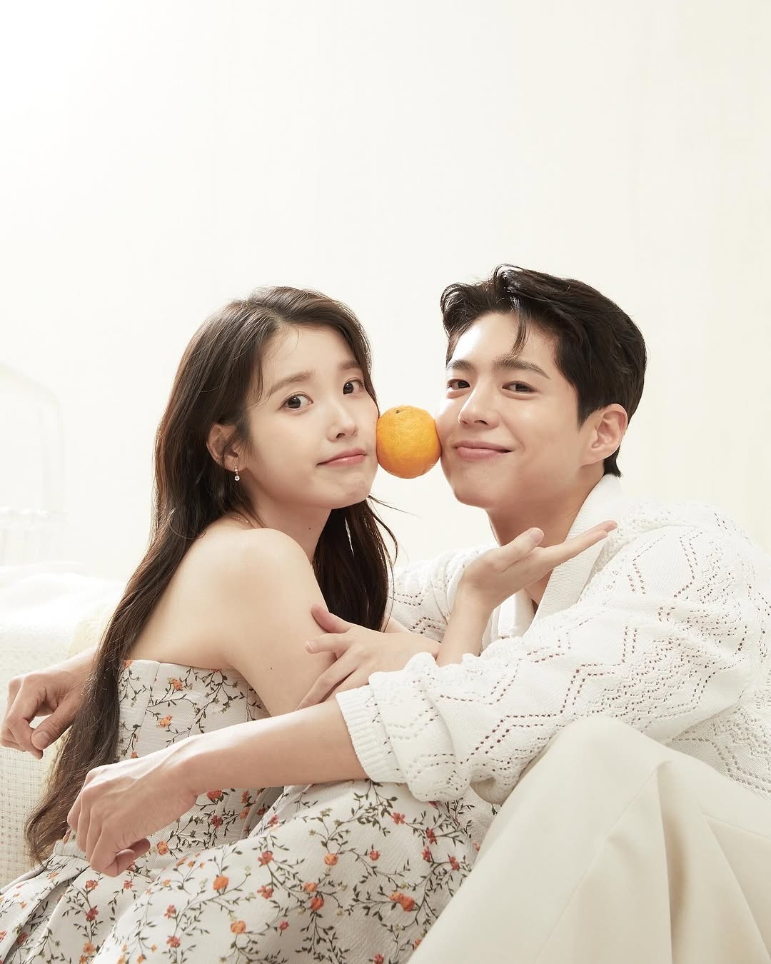 Cặp đôi IU và Park Bo Gum tạo dáng đáng yêu với quả cam giữa khuôn mặt, cùng diện trang phục trắng họa tiết hoa, gợi cảm hứng cho phong cách trang điểm tông cam tangerine rạng rỡ