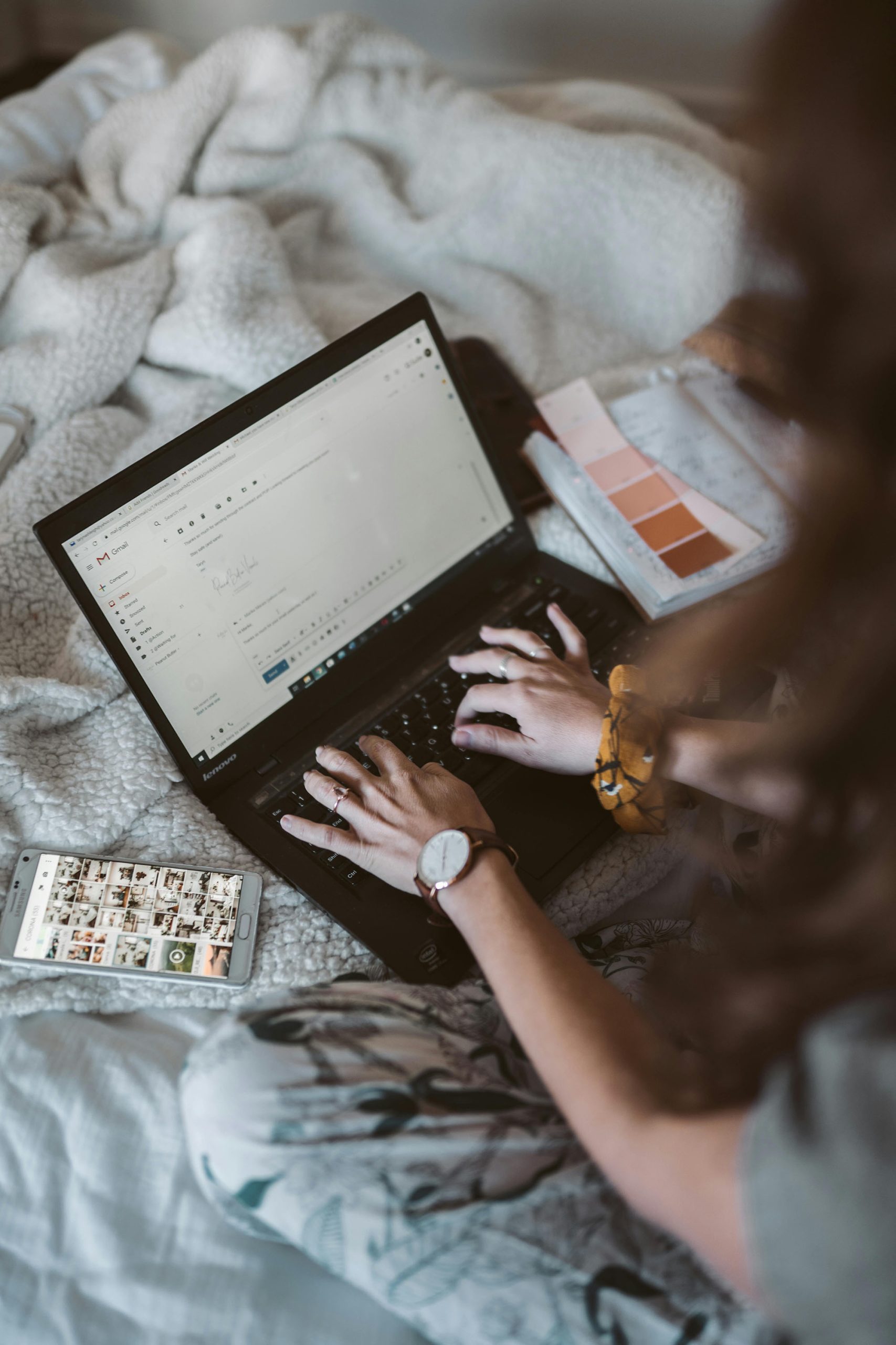 cô gái ngồi trên giường check mail trên máy tính bên cạnh điện thoại và bảng màu